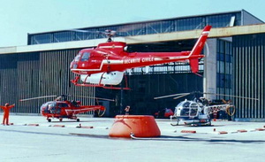 Base Hélicoptère Sécurité civile de Cannes (Ecureuil 06 en stationnaire pour le remplissage d'un réservoir d'eau lors d'une mission feu HBE) avec au sol l'Alouette III Sécurité civile "Dragon 06" et un SA315B Lama équipé HBE (F-BSUI d'Heli Union possible) au parking devant le hangar - Photo DR Chronique d'histoire - SDIS 06
